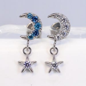 May include: A pair of silver-toned earrings. One earring features a crescent moon design with blue gemstones, and the other has white gemstones. Each moon suspends a star-shaped charm with a single gemstone.
