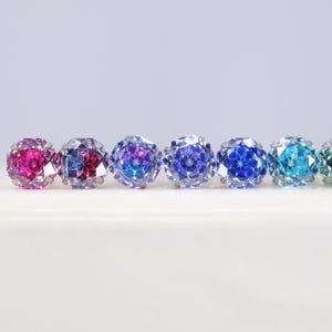 Può includere: Una fila di sette orecchini a lobo rotondi e sfaccettati, montati su griffe in metallo color argento. Le pietre presentano una sfumatura di colori, dal magenta e viola vivaci al blu intenso, ciano brillante e un tocco di verde a destra.