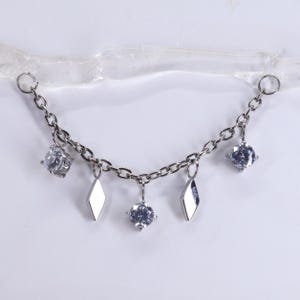 Puede incluir: Una cadena de cuerpo plateada con un diseño de cadena delicado. Cuenta con gemas claras brillantes y dijes en forma de diamante. La cadena tiene dos bucles para la fijación y está diseñada para usarse sobre el cuerpo.