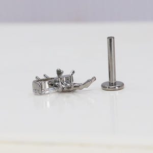 May include: A silver body jewelry piece with a clear crystal design. The jewelry is a labret stud with a flat back and a screw-on top. The crystal design is a cluster of small stones.