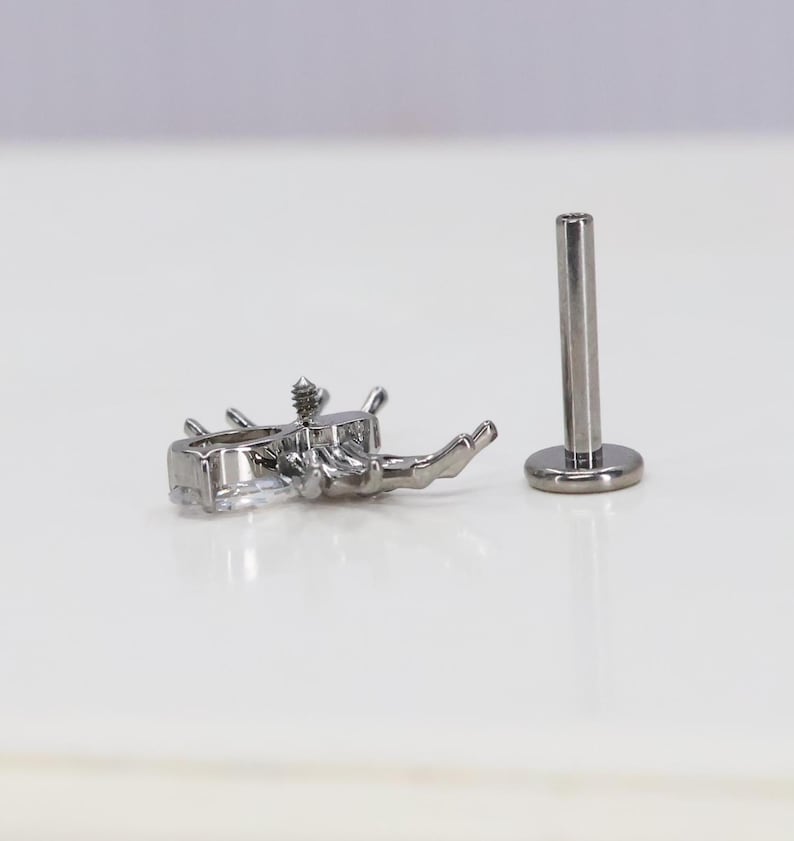 May include: A silver body jewelry piece with a clear crystal design. The jewelry is a labret stud with a flat back and a screw-on top. The crystal design is a cluster of small stones.