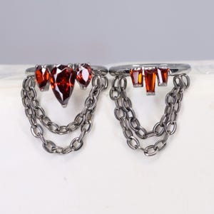 Pode incluir: Dois anéis de prata com detalhes de corrente pendurada. Um anel apresenta três pedras preciosas vermelhas em forma de gota, enquanto o outro tem quatro pedras preciosas vermelhas retangulares. As correntes são feitas de pequenas peças de prata conectadas.