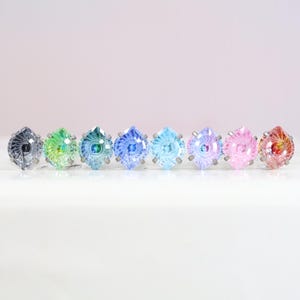 May include: A row of eight faceted, diamond-shaped stud earrings in a spectrum of colors including grey, green, teal, blue, light blue, purple, pink, and orange, each set in a silver-toned metal prong setting with a spiral pattern design.
