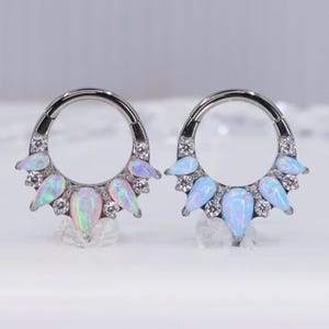 May include: Two silver septum rings, each adorned with opalescent teardrop-shaped gems and clear crystals. One ring features pink and green opals, the other blue opals. The jewelry is displayed on a white surface.