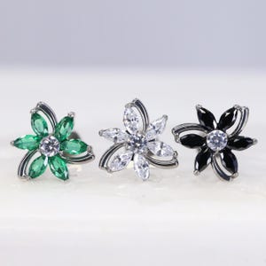 May include: Three flower-shaped stud earrings featuring marquise-cut stones arranged as petals around a central round stone. The left earring has green stones, the centre has clear stones, and the right earring has black stones, all set in silver.