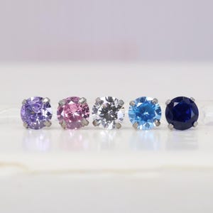 May include: Five stud earrings with round, faceted gemstones in a row. The colours are purple, pink, clear, light blue, and dark blue. Each gemstone is set in a silver-coloured metal setting.