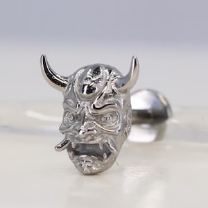 Può includere: Orecchino a lobo color argento con un design dettagliato di maschera oni. La maschera presenta corna ricurve, una bocca larga e tratti del viso intricati. Il perno dell'orecchino è visibile sul retro.