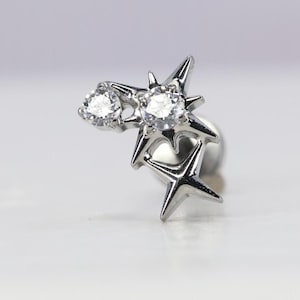 May include: A silver-toned earring featuring a starburst design. The earring has two clear, round gemstones and multiple star-shaped accents. The design is a cluster of stars, with a post for piercing.