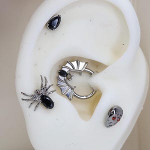 May include: A set of four black and silver body jewelry pieces. The pieces include a spider with black gems, a bat-shaped hoop with a black gem, a teardrop-shaped gem, and a skull-shaped gem with red accents.