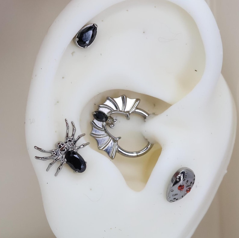 May include: A set of four black and silver body jewelry pieces. The pieces include a spider with black gems, a bat-shaped hoop with a black gem, a teardrop-shaped gem, and a skull-shaped gem with red accents.
