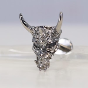 Może przedstawiać: Srebrny kolczyk z szczegółowym wzorem maski oni. Kolczyk ma zakrzywione rogi i skomplikowane rysy twarzy. Projekt jest bardzo szczegółowy i teksturowany.