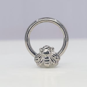 May include: Silver body jewelry featuring a bee design. The ring is a seamless hoop with a hinged closure.