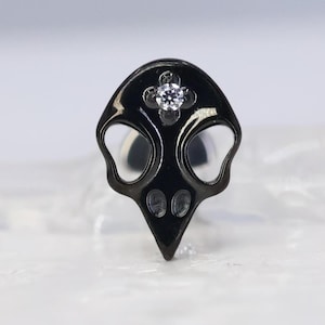 Op de afbeelding: Een zwarte, schedelvormige oorbel met een kleine, heldere edelsteen in het midden. De oorbel heeft een gepolijste afwerking en een uniek, gotisch ontwerp. De schedel heeft oog- en neusdetails.