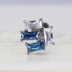 May include: A silver-toned stud earring featuring a cluster of four rectangular gemstones. Two are clear, two are a deep blue. The gemstones are set in a square arrangement, creating a modern and elegant design. The earring is suitable for ear piercings.