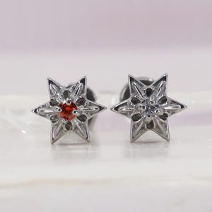 May include: A pair of silver star-shaped stud earrings. One earring has a small, round, orange gemstone at the centre, the other a clear gemstone. The earrings have a detailed, textured design.