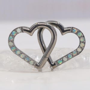 May include: A silver-colored, interlocking heart-shaped body jewelry piece. Each heart is outlined with small, iridescent, opal-like stones. The two hearts are connected, forming a single piece.