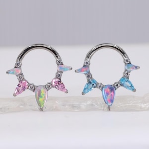 May include: Two silver-colored nose rings, each with a circular design. One ring features pink and opal-colored gemstones, while the other has blue and opal-colored gemstones. The rings are designed with a teardrop-shaped gem in the center.