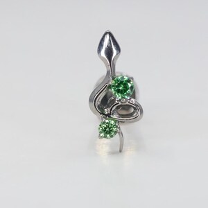 Pode incluir: Um corpo de cobra prateado com uma gema verde na cauda e outra gema verde na cabeça. A cobra está enroscada em um design decorativo.