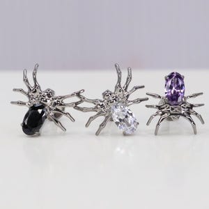 May include: Three silver spider earrings with different colored gemstones. The first earring has a black gemstone, the second earring has a clear gemstone, and the third earring has a purple gemstone.