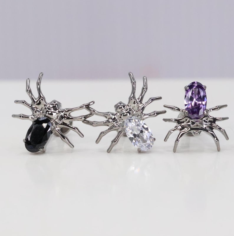 May include: Three silver spider earrings with different colored gemstones. The first earring has a black gemstone, the second earring has a clear gemstone, and the third earring has a purple gemstone.