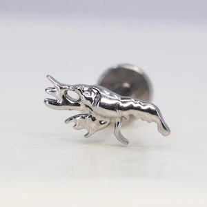May include: A silver-coloured earring stud in the shape of a crawfish. The crawfish has detailed features, including claws, antennae, and a segmented body. The earring post is visible behind the crawfish.