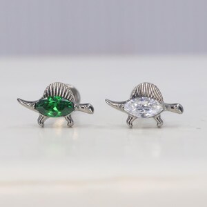 May include: Two silver-toned dinosaur stud earrings. One earring features a green gemstone, while the other has a clear, white gemstone. The earrings are set against a light background.
