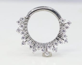 Puede incluir: Un anillo de plata con un diseño circular que presenta una fila de pequeñas piedras transparentes alrededor del borde. El anillo está adornado con un grupo de piedras más pequeñas que se extienden hacia afuera desde el centro.