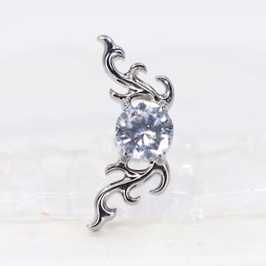 May include: A silver-toned metal body jewellery piece featuring a central round clear gemstone set within a curved, ornate frame with scrolling filigree details. The design is symmetrical, with intricate metalwork extending above and below the stone.