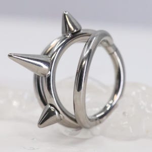 May include: A silver-colored ring with three conical spikes. The ring has a double-band design, with one band wrapped around the other. The spikes are evenly spaced around the outer band. The ring is made of a shiny, metallic material.