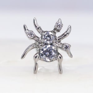 May include: A silver-toned spider-shaped jewellery piece featuring a polished metallic finish. The body and legs are adorned with multiple clear, round-cut faceted stones that catch the light, creating a sparkling effect across the entire design.