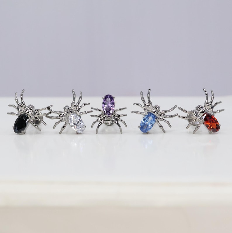 May include: Five silver spider earrings with different colored gemstones. The gemstones are black, white, purple, blue, and red.