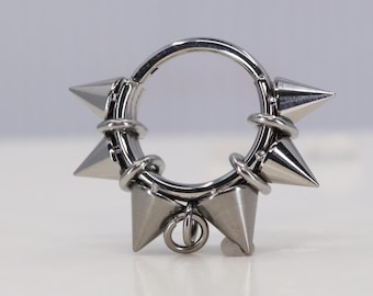 May include: A silver-coloured, hinged piercing ring featuring six conical spikes evenly spaced around the circumference. The ring has a small, dangling loop at the bottom. The spikes are pointed and metallic.