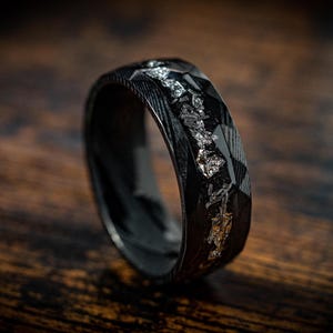 May include: A black ring with a textured finish and a silver inlay. The ring is made of carbon fiber and has a unique design.