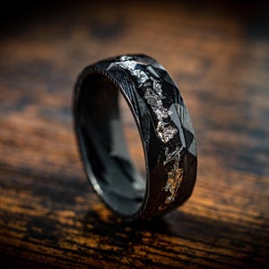 May include: A black and silver ring with a textured finish. The ring has a band of silver metal embedded in the black metal.