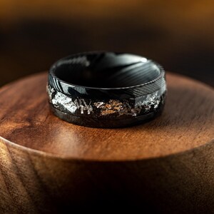 May include: A black and silver ring with a textured finish. The ring is on a wooden display stand.