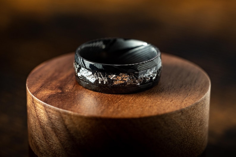 May include: A black and silver ring with a textured finish. The ring is on a wooden display stand.
