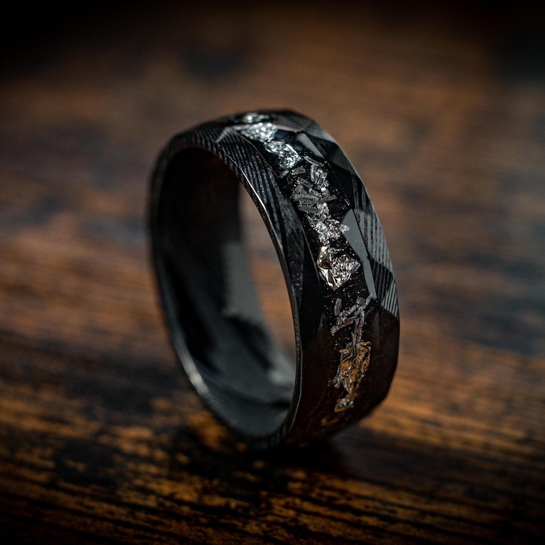May include: A black ring with a textured finish and a silver inlay. The ring is made of carbon fiber and has a unique design.
