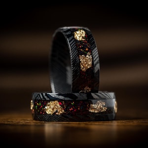 May include: Two black rings with a Damascus steel pattern and inlaid with red and gold flakes. The rings are stacked on top of each other, showcasing the intricate design and craftsmanship. The background is a dark brown.