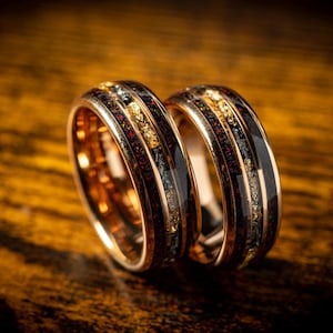 May include: Two matching rose gold wedding bands with black and red inlay and gold flecks.