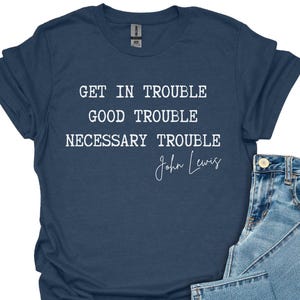 May include: A navy blue t-shirt with the quote "GET IN TROUBLE GOOD TROUBLE NECESSARY TROUBLE" and the signature "John Lewis" in white. The shirt is paired with light blue denim jeans.