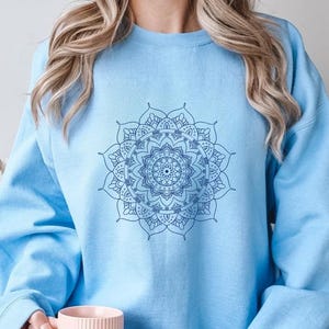 May include: A light blue sweatshirt with a blue mandala design on the front. The sweatshirt is being worn by a person holding a light pink mug.