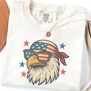 May include: An off-white t-shirt featuring a graphic of a bald eagle wearing a bandana and sunglasses, both patterned with the American flag. The design is surrounded by small red and blue stars. The shirt has a Comfort Colors brand neck label.