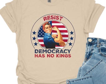 May include: A beige t-shirt with a graphic of a woman flexing her arm, set against a US flag design. The text "RESIST" and "DEMOCRACY HAS NO KINGS" are displayed. Light blue denim jeans are folded next to the shirt.