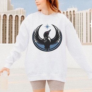 May include: White sweatshirt featuring a black and blue graphic of a bird with outstretched wings, a star, and a circular design. The word "white" is in a white starburst shape. The sweatshirt is a long-sleeved, crew neck style.