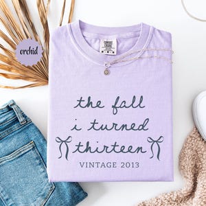 Puede incluir: Una camiseta de color morado claro con texto en gris oscuro que dice the fall i turned thirteen VINTAGE 2013, con un pequeño gráfico de lazo a cada lado de la palabra thirteen. La camiseta tiene cuello redondo y una etiqueta Comfort Colors.