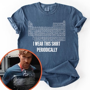 May include: Denim blue t-shirt featuring a white periodic table design and the phrase "I Wear This Shirt Periodically." The shirt has rolled-up sleeves and a classic crew neck. A person wearing a similar shirt is in the image.