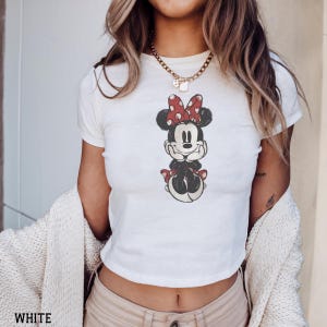 May include: White crop top featuring a Minnie Mouse graphic. The graphic shows Minnie Mouse with a red bow with white polka dots. The word "WHITE" is printed at the bottom of the image.