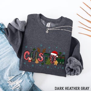 May include: Dark heather gray sweatshirt with the word "CUSTOM" in a Christmas-themed design. The letters are decorated with plaid patterns, a Santa hat, snowflakes, and gingerbread men. A silver necklace is draped across the sweatshirt. The text "DARK HEATHER GRAY" is at the bottom.