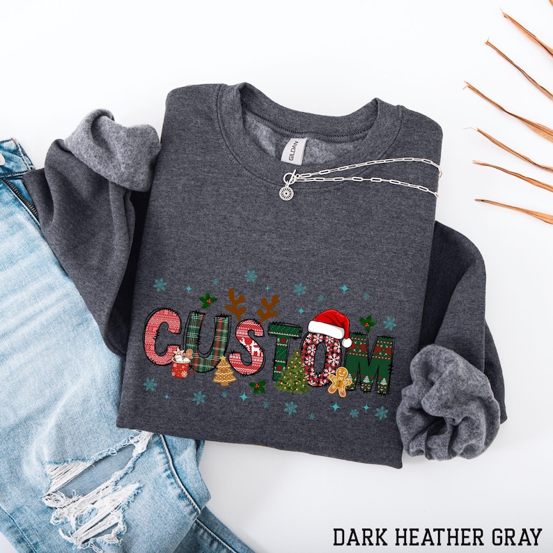 May include: Dark heather gray sweatshirt with the word "CUSTOM" in a Christmas-themed design. The letters are decorated with plaid patterns, a Santa hat, snowflakes, and gingerbread men. A silver necklace is draped across the sweatshirt. The text "DARK HEATHER GRAY" is at the bottom.