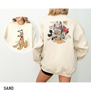 May include: Sand-colored sweatshirt featuring a Pluto graphic on the front and a group of Disney characters on the back. The Pluto graphic shows the dog in a seated position. The back of the sweatshirt features Mickey Mouse, Minnie Mouse, Donald Duck, Daisy Duck, Goofy, and Pluto.